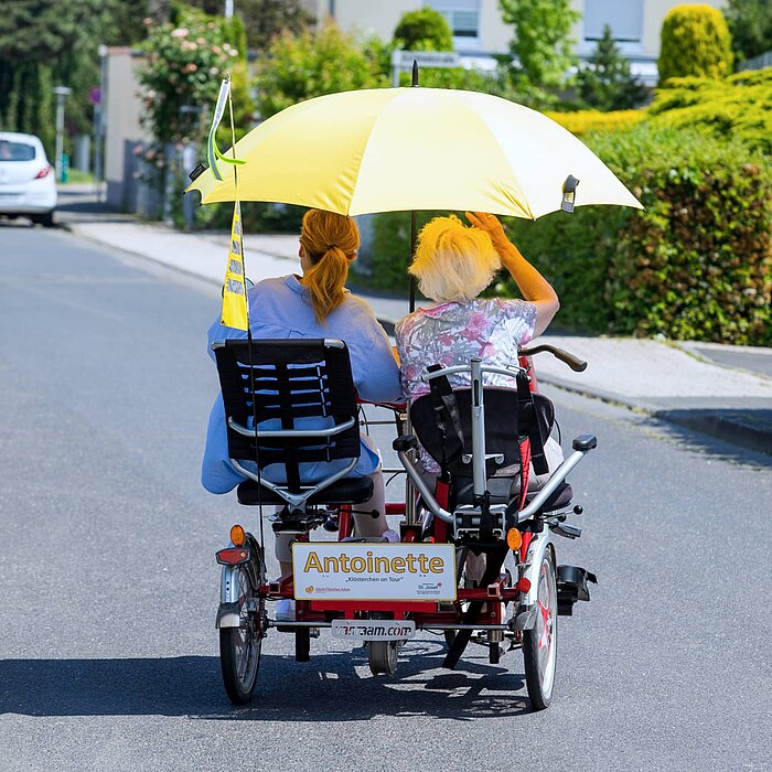 Eine ältere Dame fährt mit ihrer Begleiterin auf einem Dreirad-Tandem auf einer Straße in Richtung Natur. Da die Sonne scheint, haben die beiden Personen einen Sonnenschirm aufgespannt.