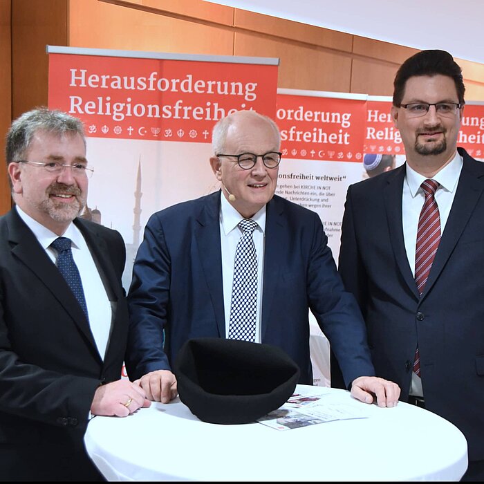 Das Bild zeigt Volker Kauder, Christoph Konopka und Mathias Junggeburth. Alle drei Herren tragen eine Brille, lächeln in die Kamera und schauen nach rechts. Im Hintergrund sind Transparente zu sehen, die auf das Thema Religionsfreiheit aufmerksam machen sollen.