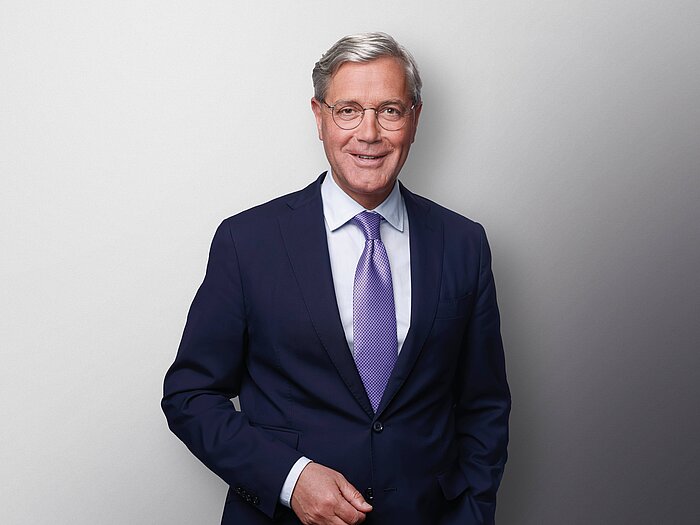 Das Bild zeigt Dr. Norbert Roettgen. Er lächelt freundlich und trägt ein dunkelblaues Jackett mit einer lila glänzenden Krawatte.