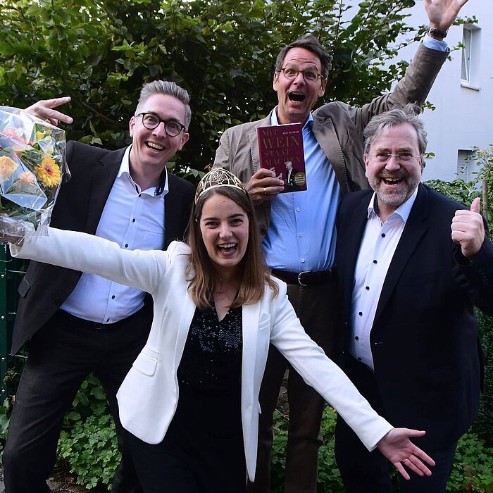 Man sieht ein Gruppenfoto mit Christoph Konopka, Dr. Knut Bergmann und Clemens Pollmann mit der 72. Deutschen Weinkönigin Eva Lanzerath. Alle vier Personen drücken ihre Freude durch Handgesten aus.